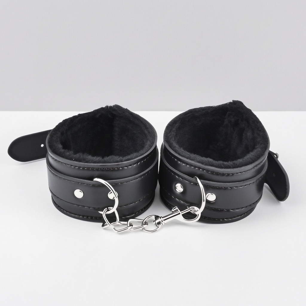 Black faux-leather ankle cuffs with plush black faux-fur lining and silver metal rings, chain, and snap hooks, displayed on a white surface against a light grey background - as part of a couples' bondage kit.