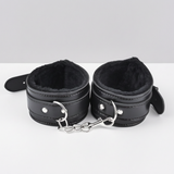 Black faux-leather ankle cuffs with plush black faux-fur lining and silver metal rings, chain, and snap hooks, displayed on a white surface against a light grey background - as part of a couples' bondage kit.